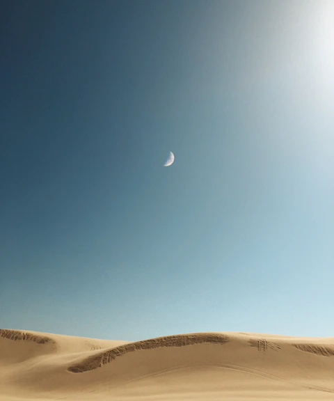 A crescent moon in a blue sky above smooth golden sand dunes